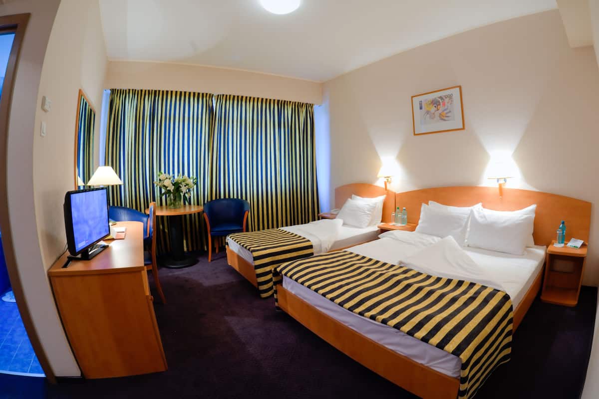 A hotel room with two beds, a desk with a computer, a chair, a small table with a vase of flowers, and a window with striped curtains.