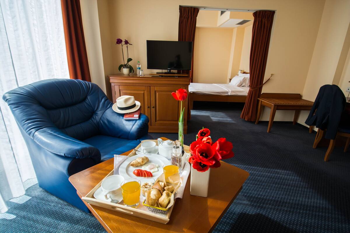 A hotel room with a blue leather sofa, a wooden coffee table with breakfast items, a TV on a wooden cabinet, and a bed with a wooden headboard.