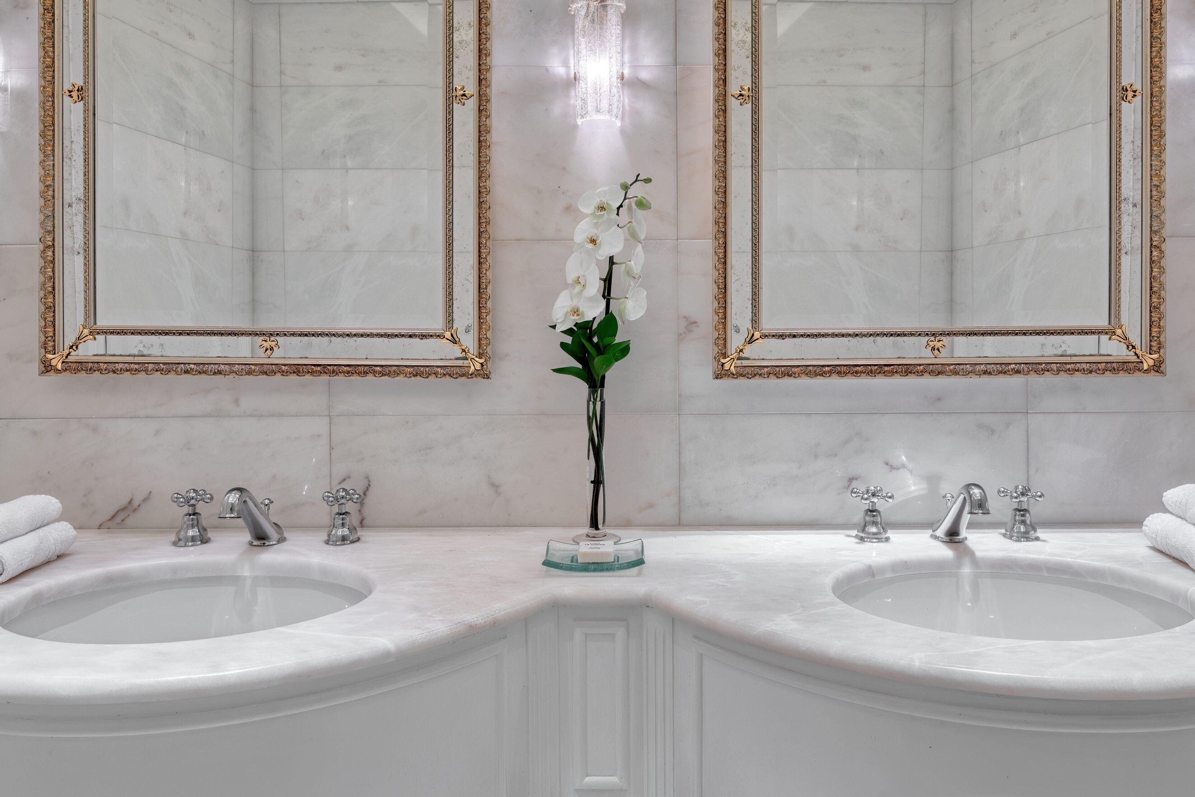 Een badkamer met twee wasbakken, een vaas met witte bloemen en een spiegel met gouden rand.