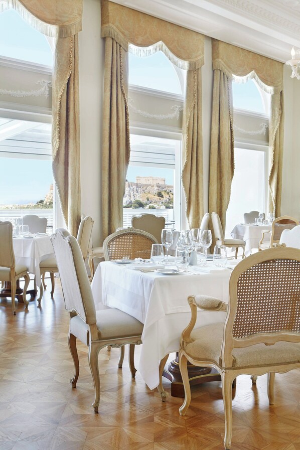 A dining area with tables set for six, elegant chairs, and large windows with curtains.