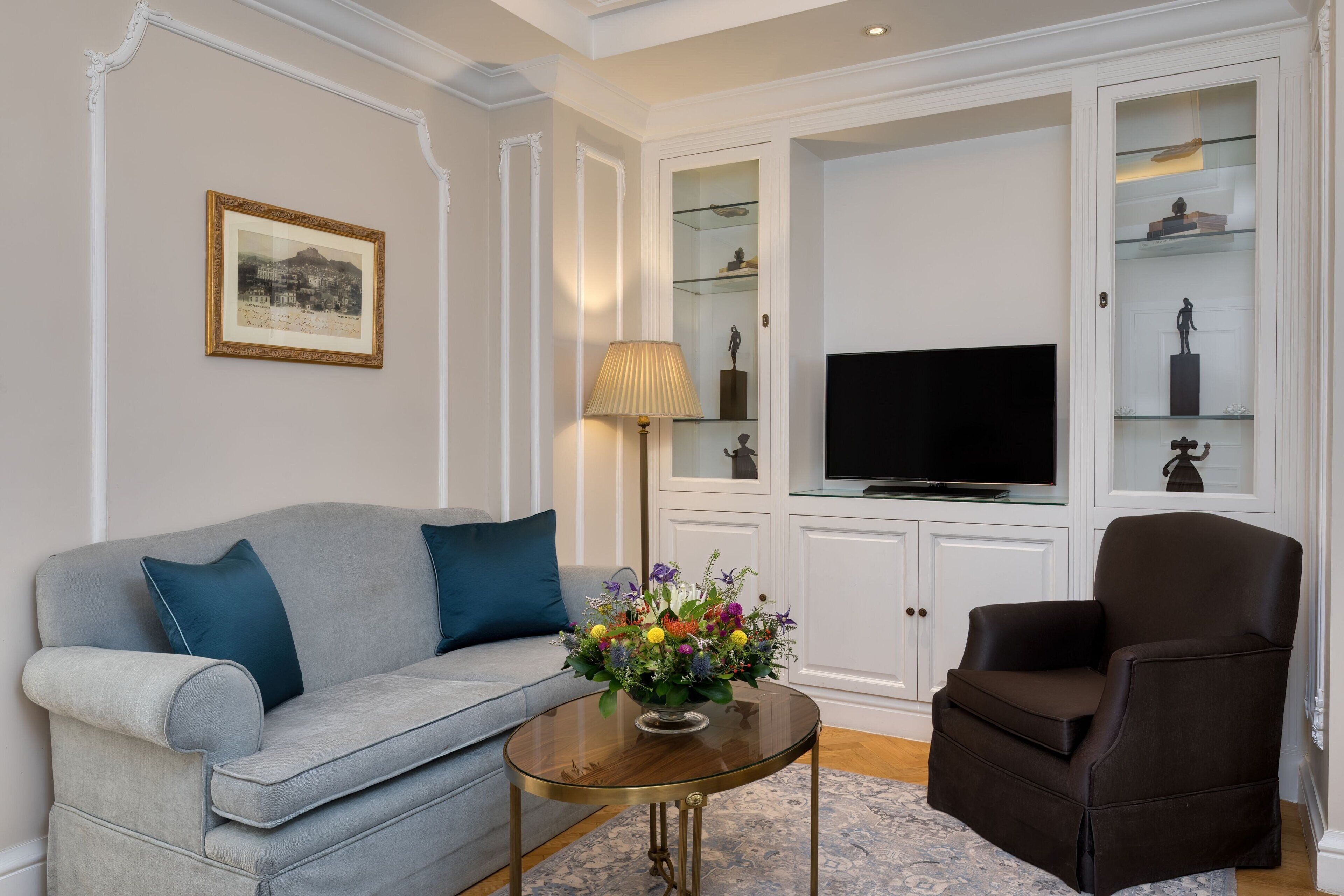 A living room with a grey sofa, a brown armchair, a glass coffee table, and a TV.