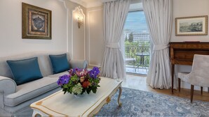 A living room with a sofa, coffee table, and piano. There is a balcony with curtains and a view of buildings. - King George, a Luxury Collection Hotel, Athens (Athens)
