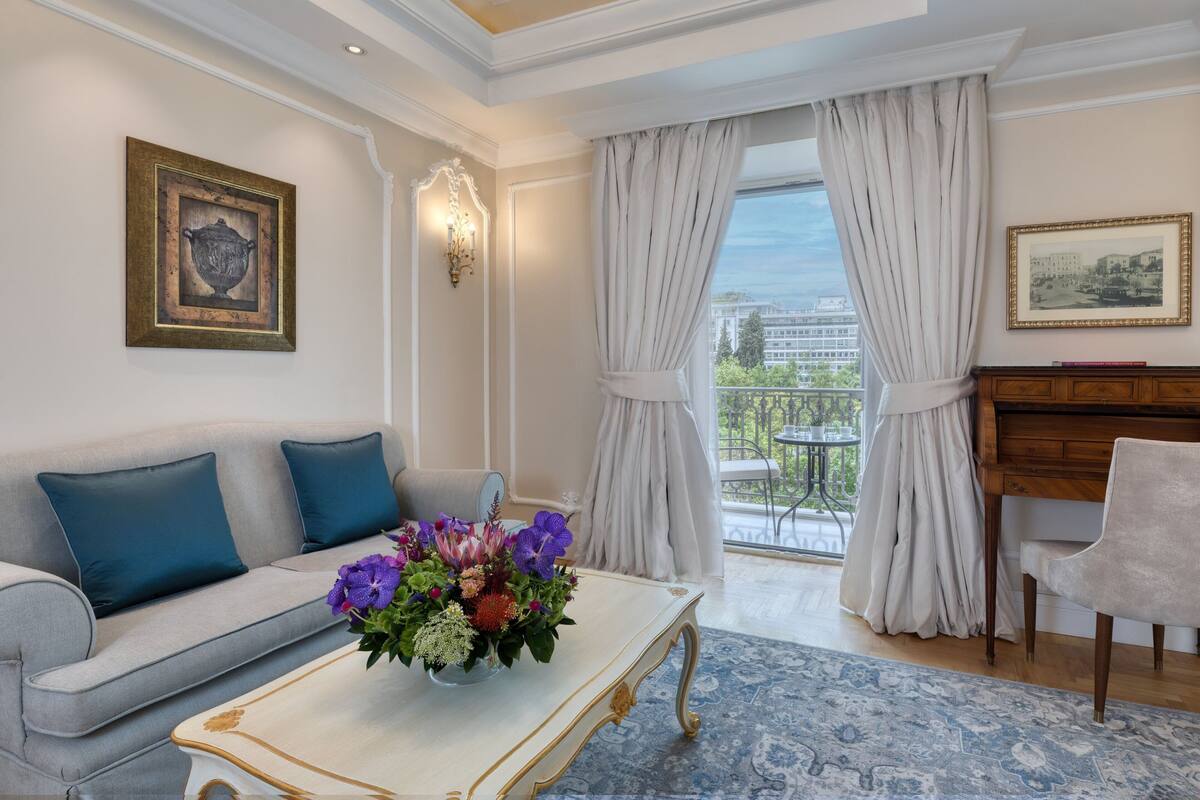 Un soggiorno con divano, tavolino da caffè e pianoforte. Presente un balcone con tende e vista sugli edifici.