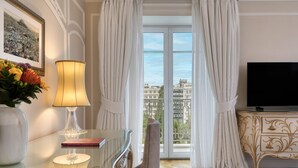 A room with a glass table, a lamp, a book, a television, and a view of buildings through large windows. - King George, a Luxury Collection Hotel, Athens (Athens)