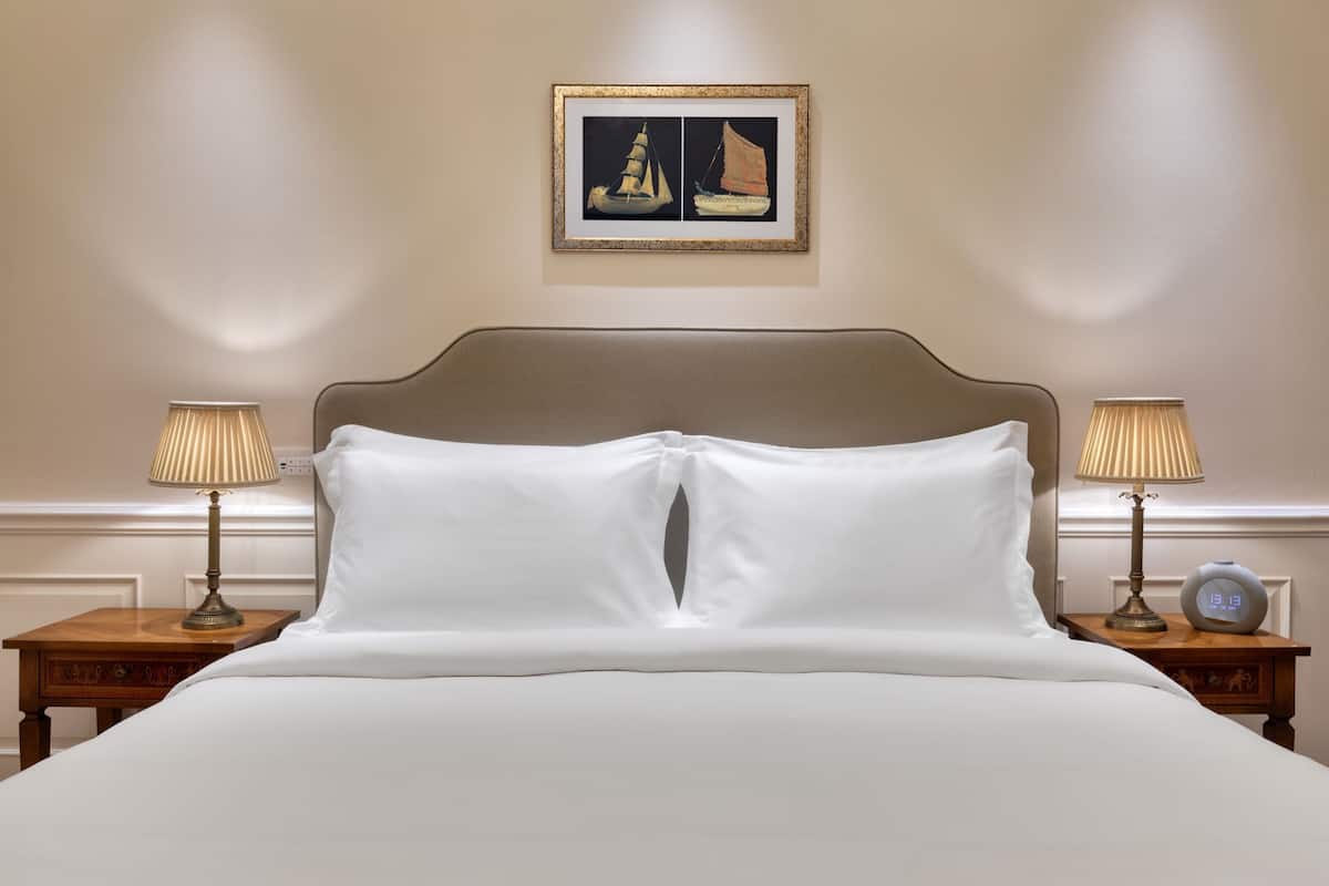Un letto rifatto con lenzuola bianche, con due comodini ai lati, ognuno con una lampada e una sveglia. Sopra il letto, un quadro con due barche a vela.