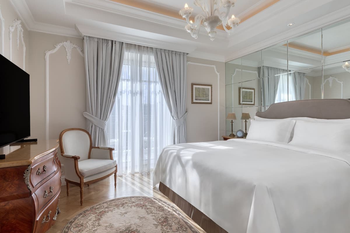 Camera d'albergo con un letto grande, una cassettiera in legno, una sedia, un televisore e un ampio specchio.