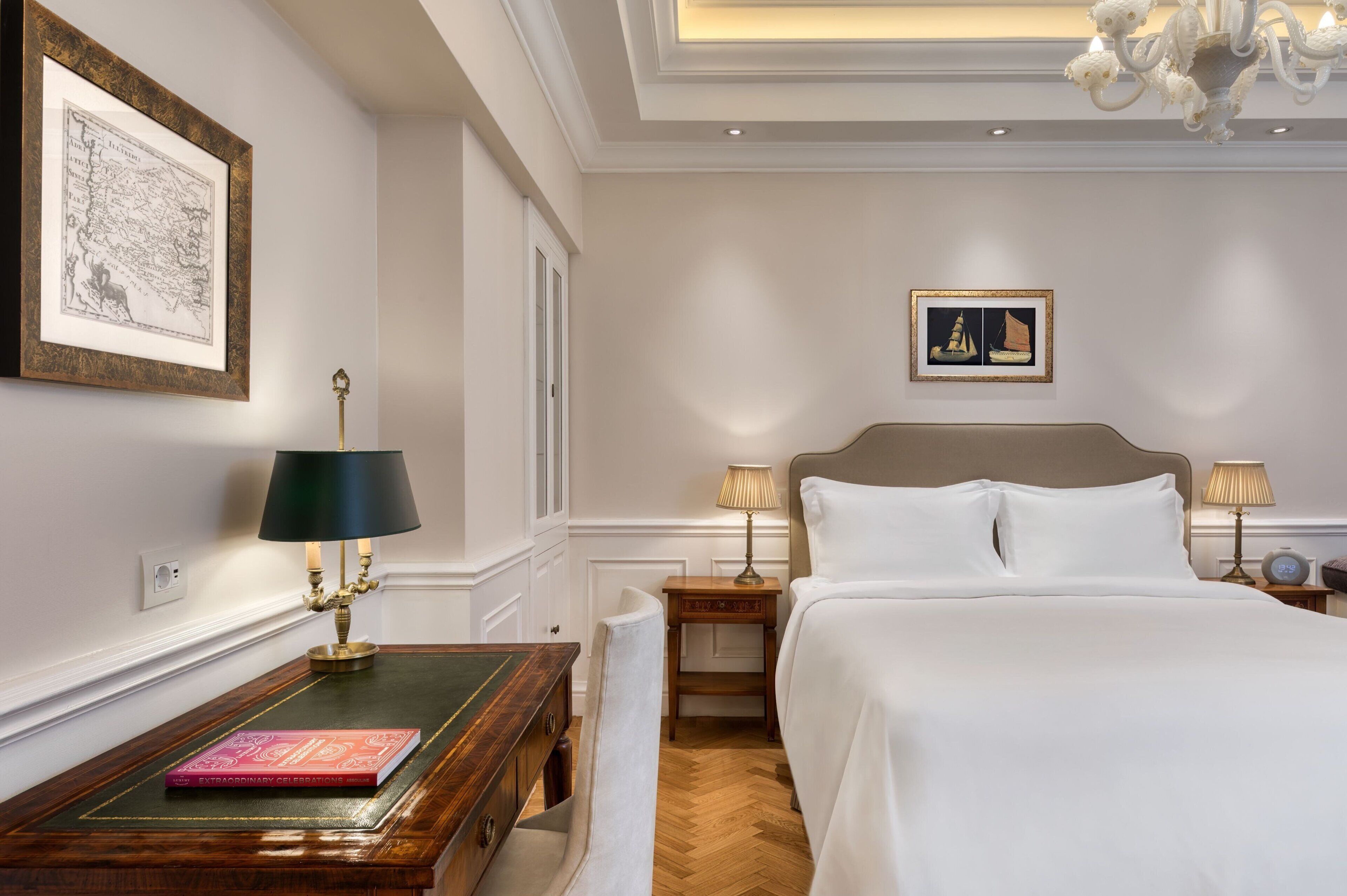 Habitación de hotel con una cama grande, una mesita de noche con lámpara, un mapa enmarcado en la pared y una estantería con un libro.