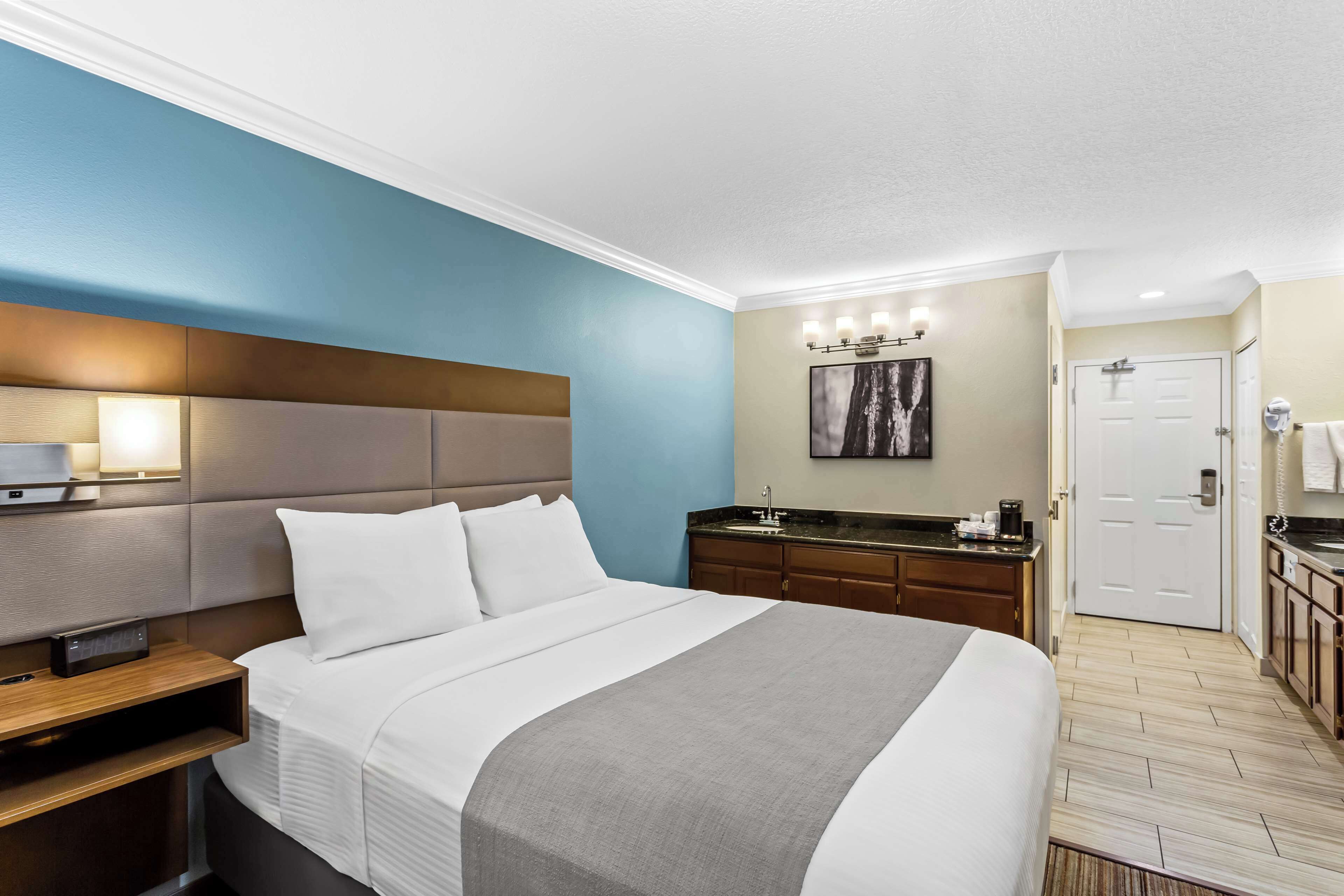 A hotel room with a large bed, a bedside table, a nightstand, a bathroom with a sink and mirror, and a door leading to another room.
