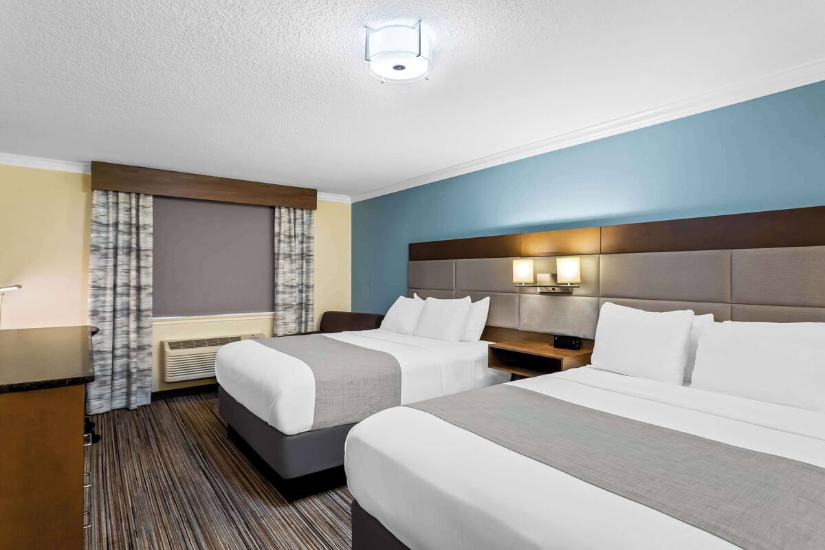 A hotel room with two beds, a desk, a chair, a window with curtains, a wall-mounted lamp, and a ceiling light.