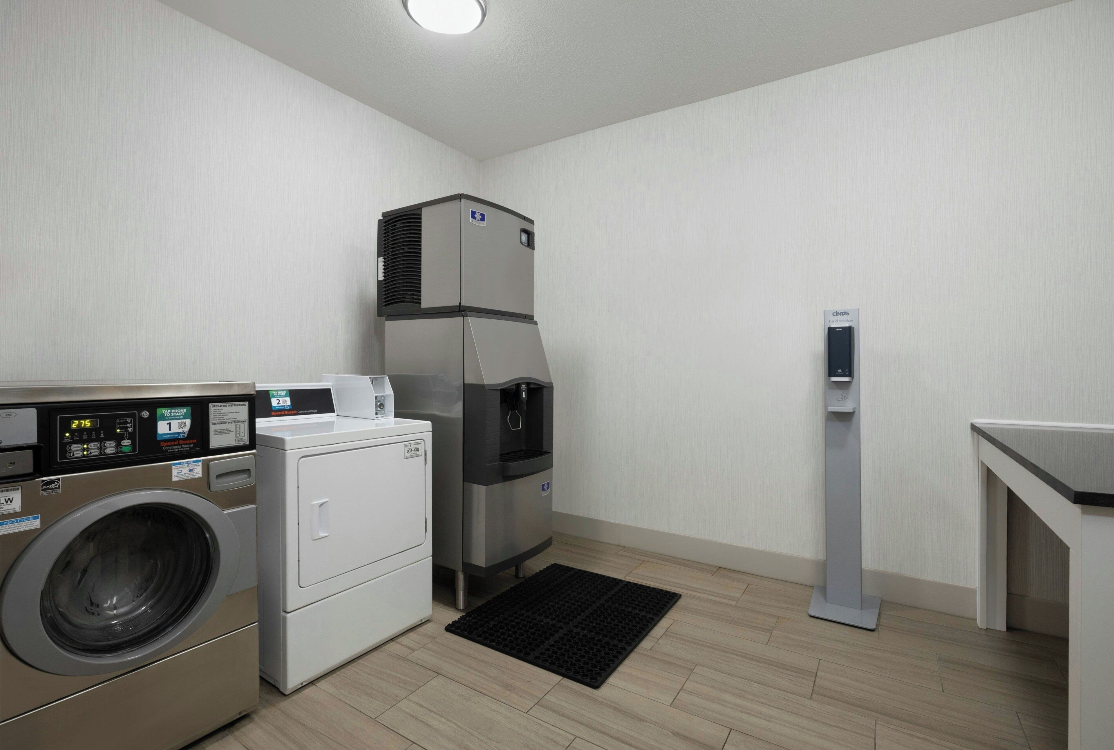 A compact hotel room with a washing machine, a compact refrigerator, and a hand sanitizer dispenser.