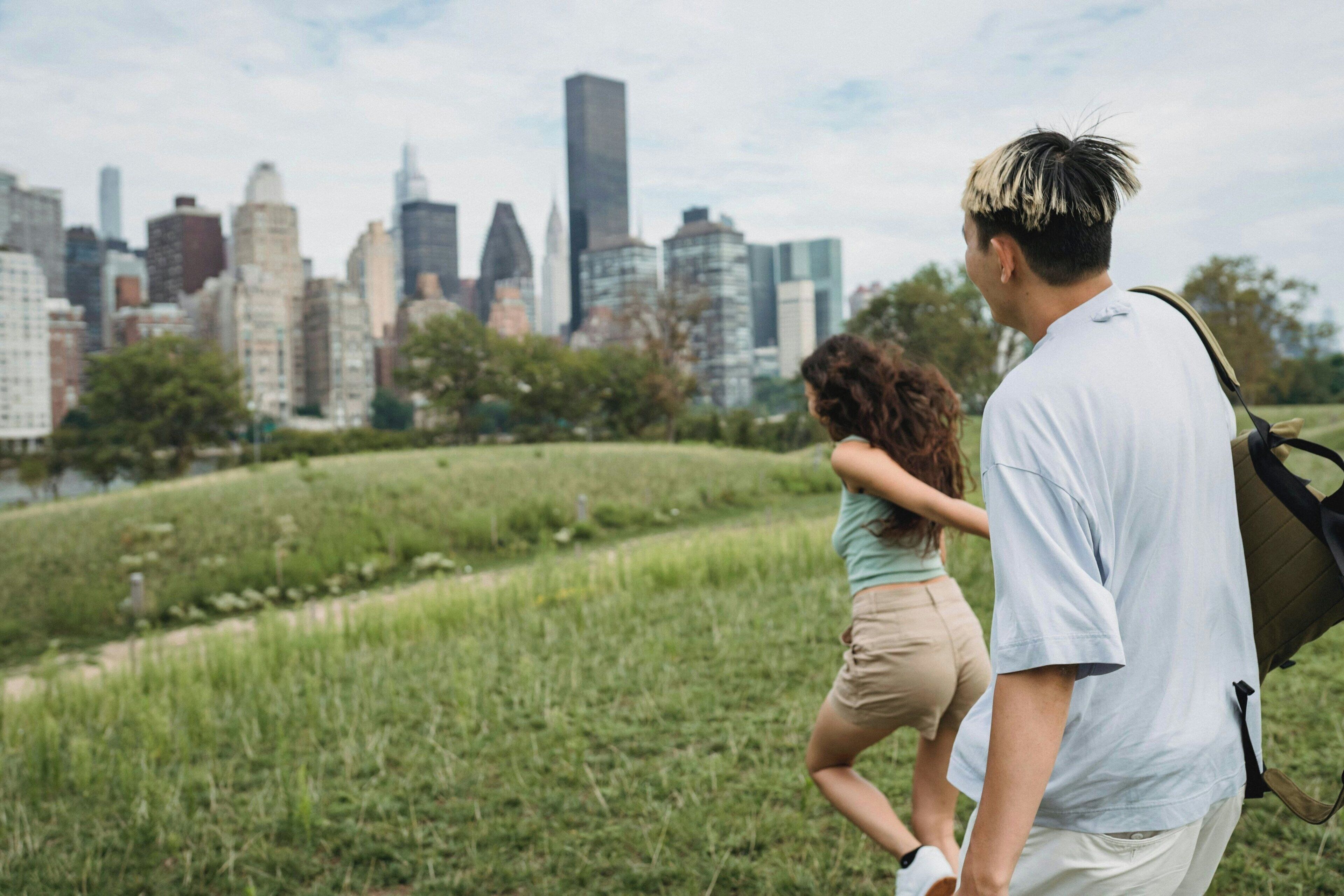 A couple walking on a grassy path with a city skyline in the background.