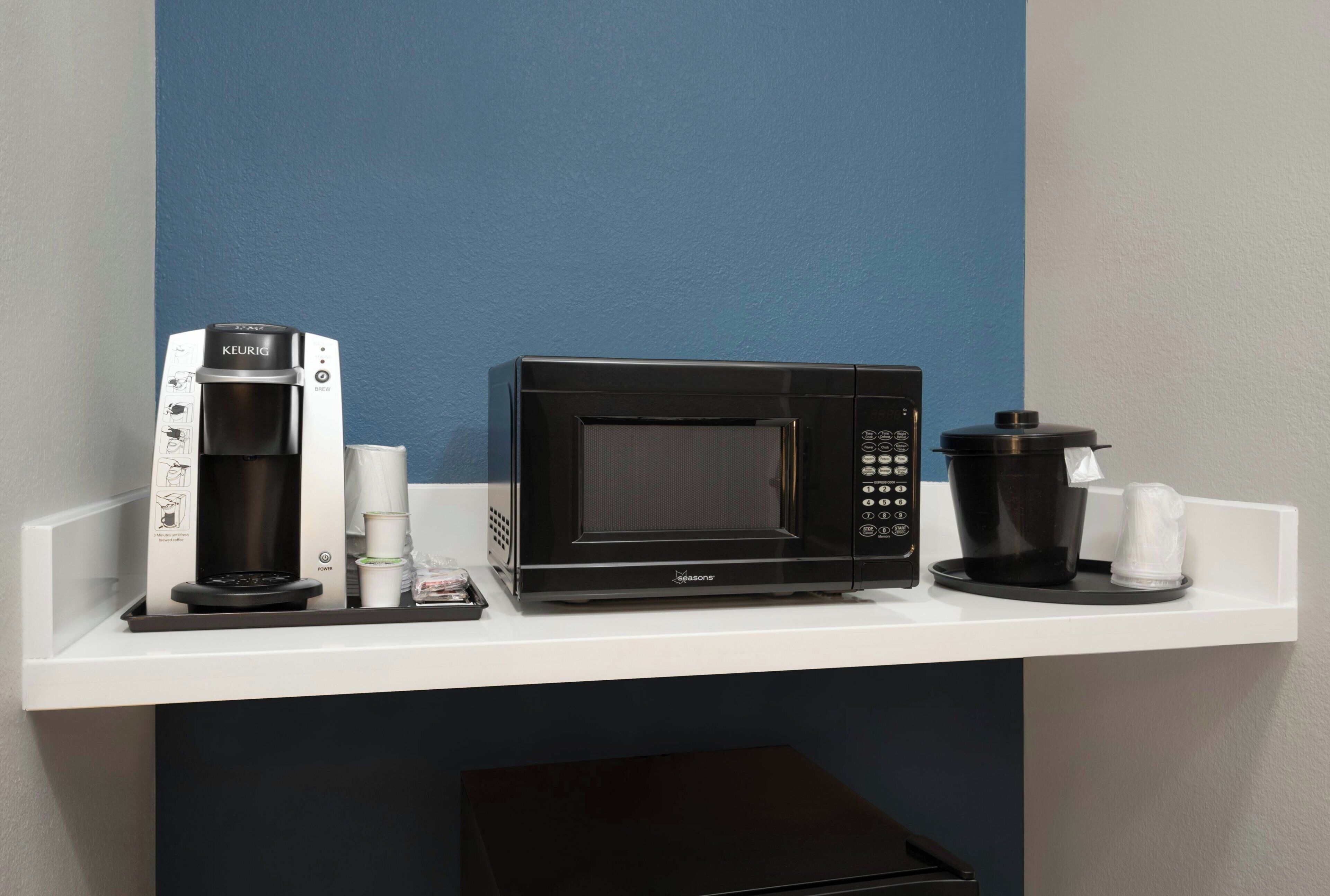 Et tekøkken med en Keurig kaffemaskine, en mikrobølgeovn og en kaffekværn på en hvid hylde.