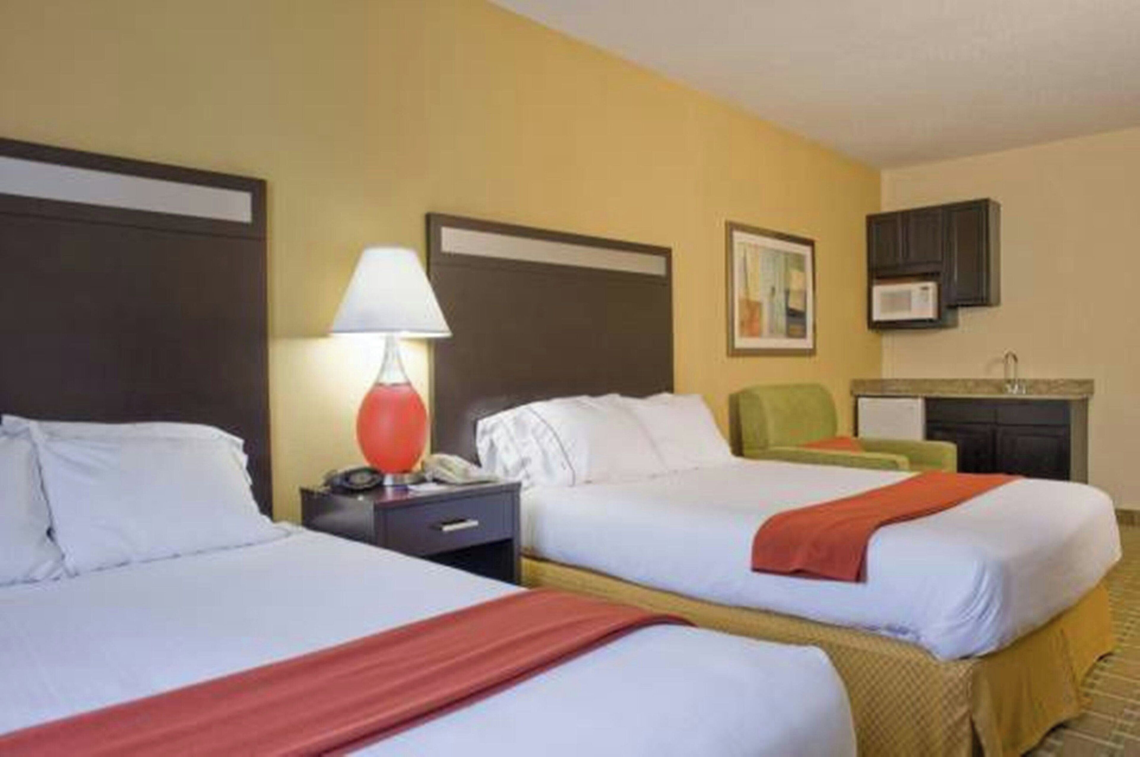 A hotel room with two beds, a nightstand, a lamp, a painting, and a kitchenette.