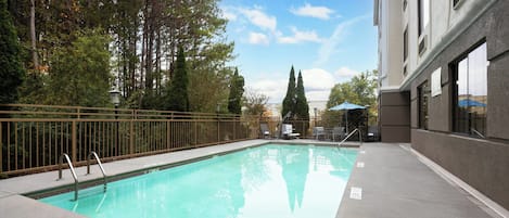 A swimming pool with a metal ladder, surrounded by a fence, trees, and a building with windows.