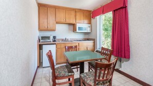 A compact kitchen with wooden cabinets, a microwave, and a small refrigerator. There is a dining table with four chairs, and a window with red curtains. - Days Inn by Wyndham Glen Allen/Richmond North (Glen Allen)
