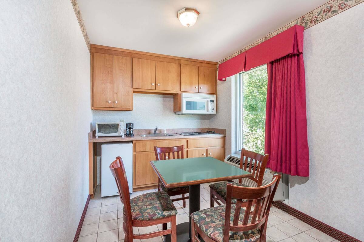 A compact kitchen with wooden cabinets, a microwave, and a small refrigerator. There is a dining table with four chairs, and a window with red curtains.