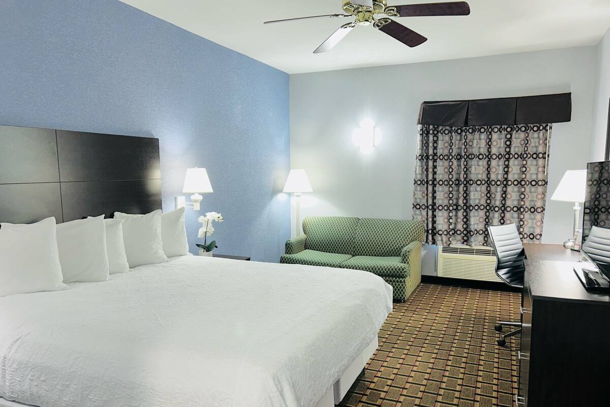 A hotel room with a large bed, a green armchair, a desk with a computer, and a ceiling fan.