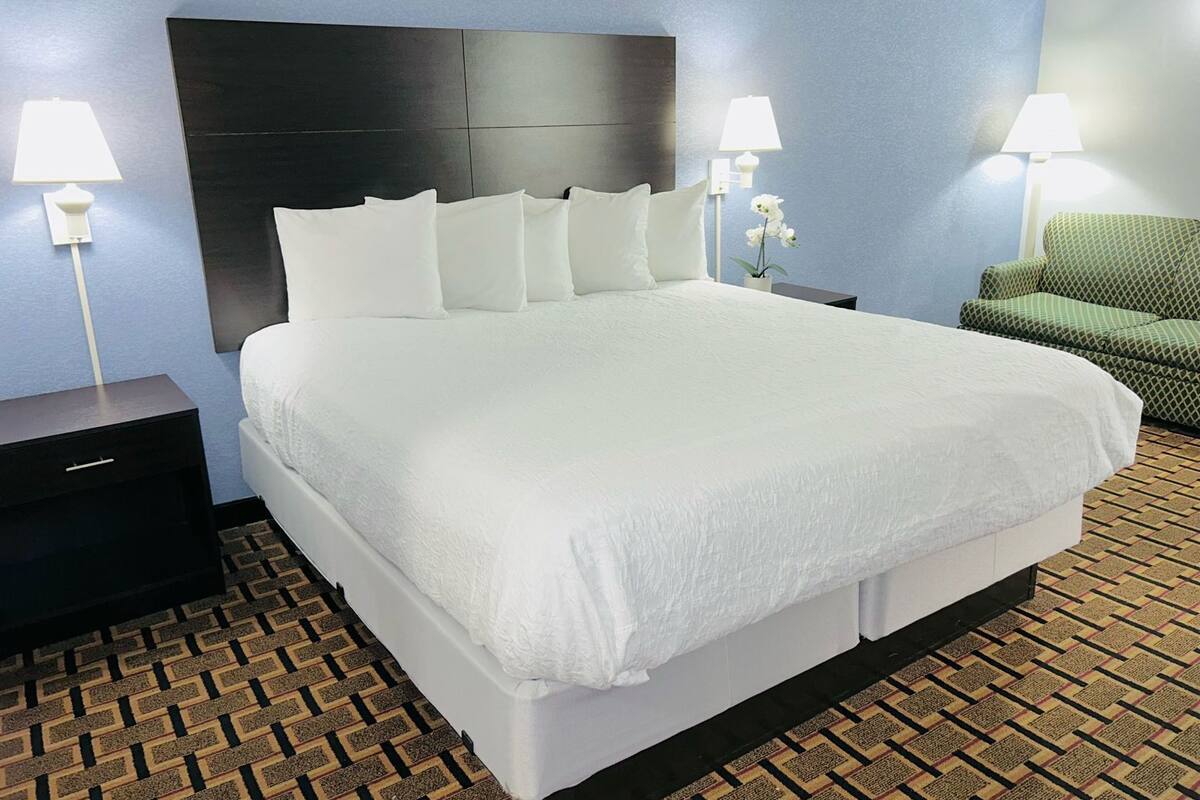 A hotel room with a large bed, two bedside tables with lamps, a green chair, and patterned carpet.
