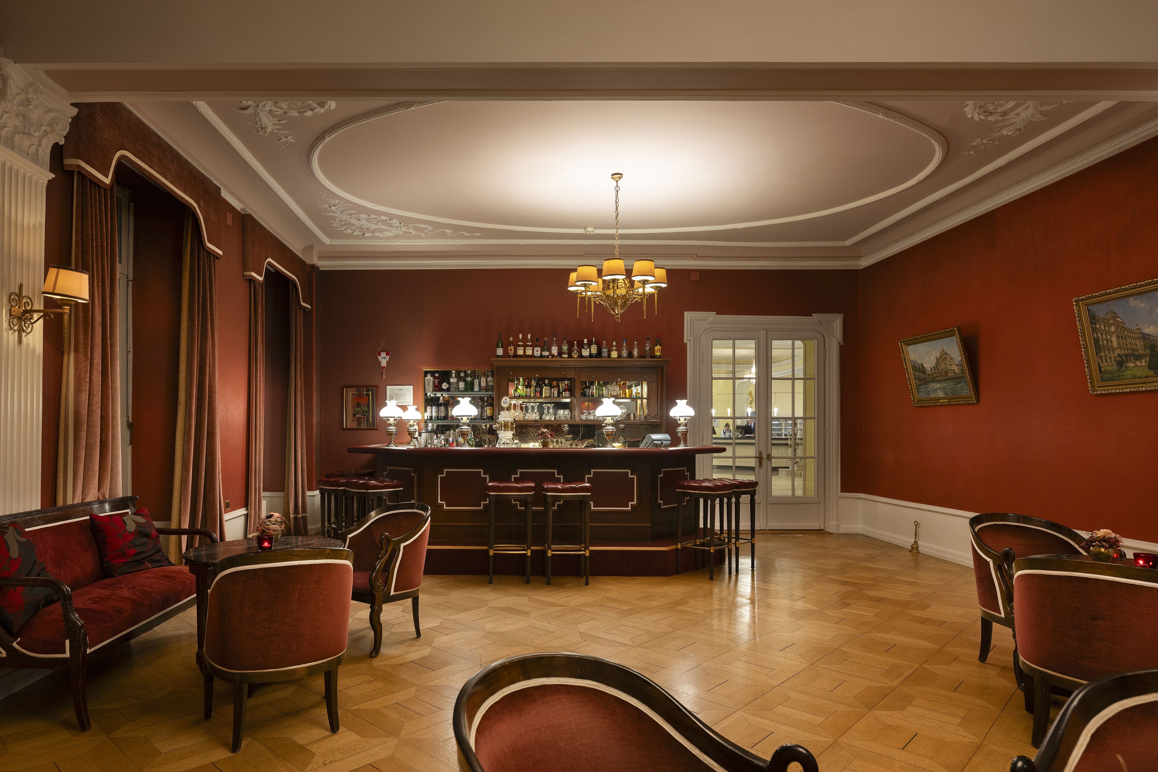 A bar area with a wooden floor, red upholstered chairs, a bar counter, and a chandelier.