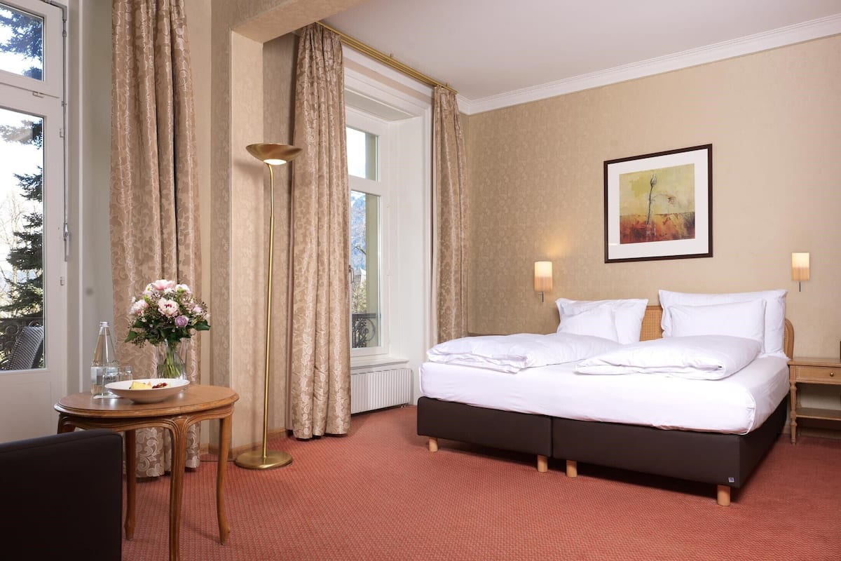 Ett hotellrum med en stor säng, ett nattduksbord med en vas med blommor, ett fönster med gardiner och en inramad tavla på väggen.