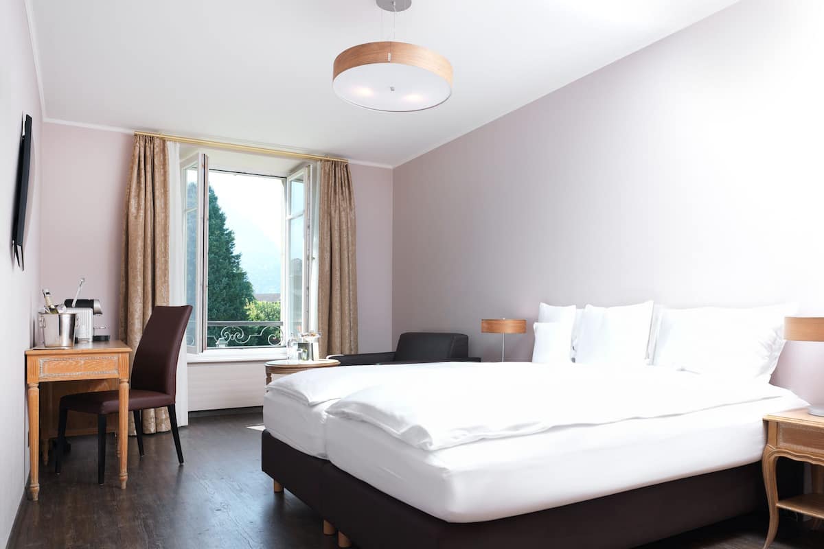 Ett hotellrum med en stor säng, ett skrivbord med en stol, ett litet bord och ett fönster med gardiner.