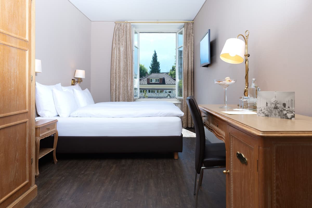 Ett hotellrum med en säng, ett skrivbord, en stol, en tv, en lampa och ett fönster med utsikt över en byggnad.