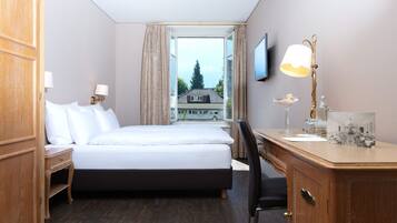 Quarto de hotel com cama, escrivaninha, cadeira, televisão, luminária e janela com vista para um edifício.