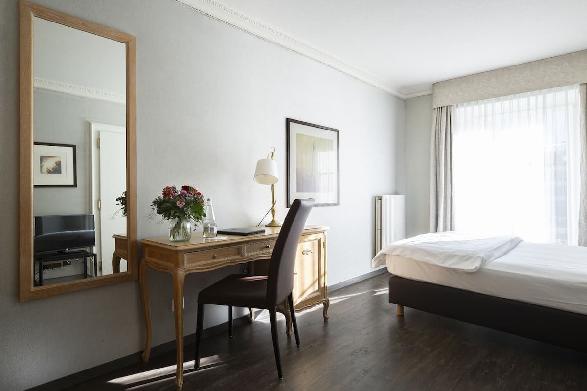 Ett hotellrum med en stor säng, ett skrivbord i trä med en stol, en skrivbordslampa, en inramad tavla och en stor spegel.