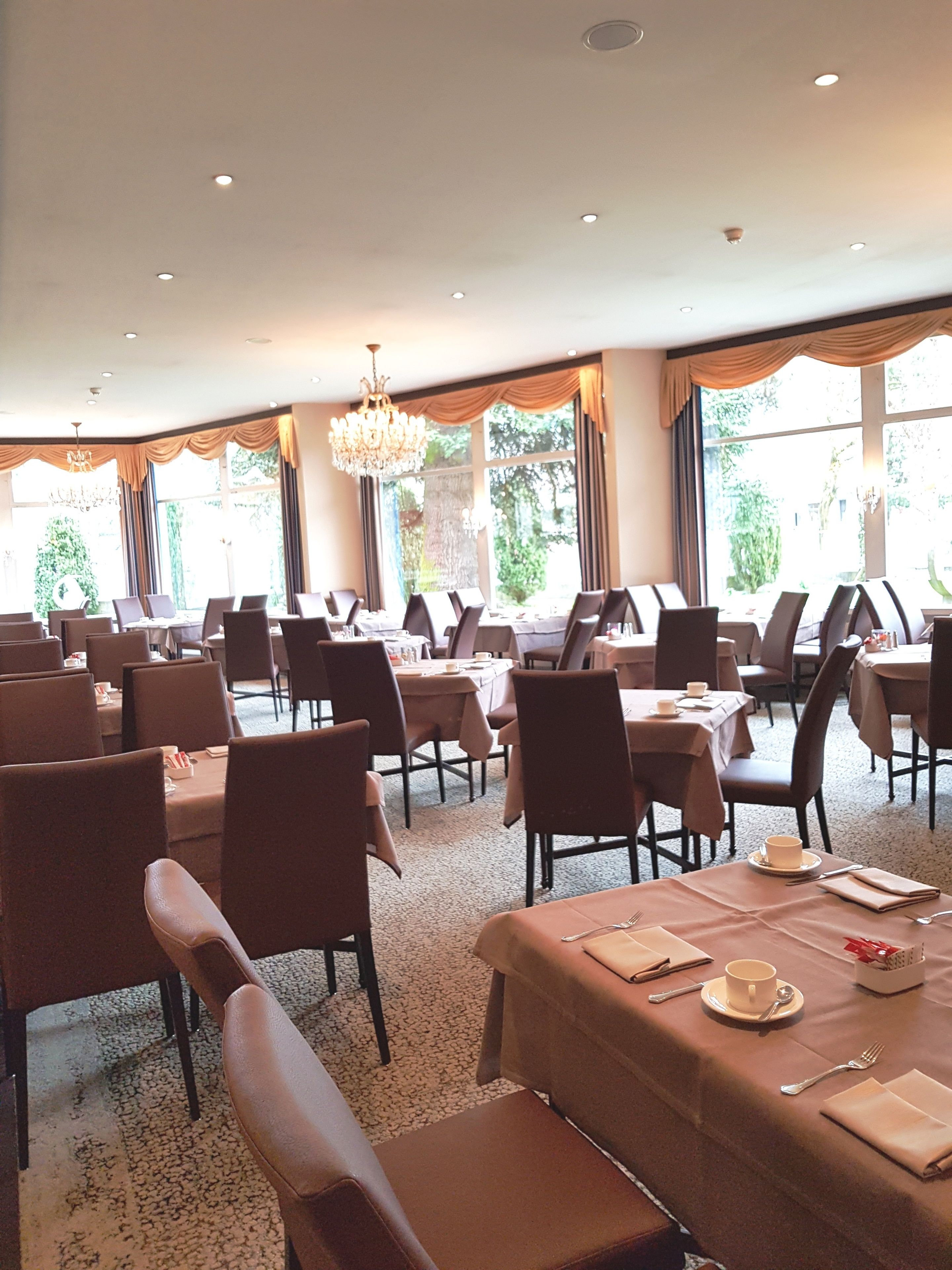 Ein geräumiger Essbereich mit mehreren Tischen und Stühlen, großen Fenstern mit Vorhängen und einer Chandelier.