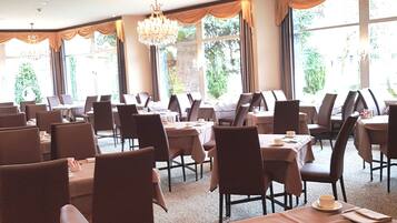 A spacious dining area with multiple tables and chairs, large windows with curtains, and a chandelier.