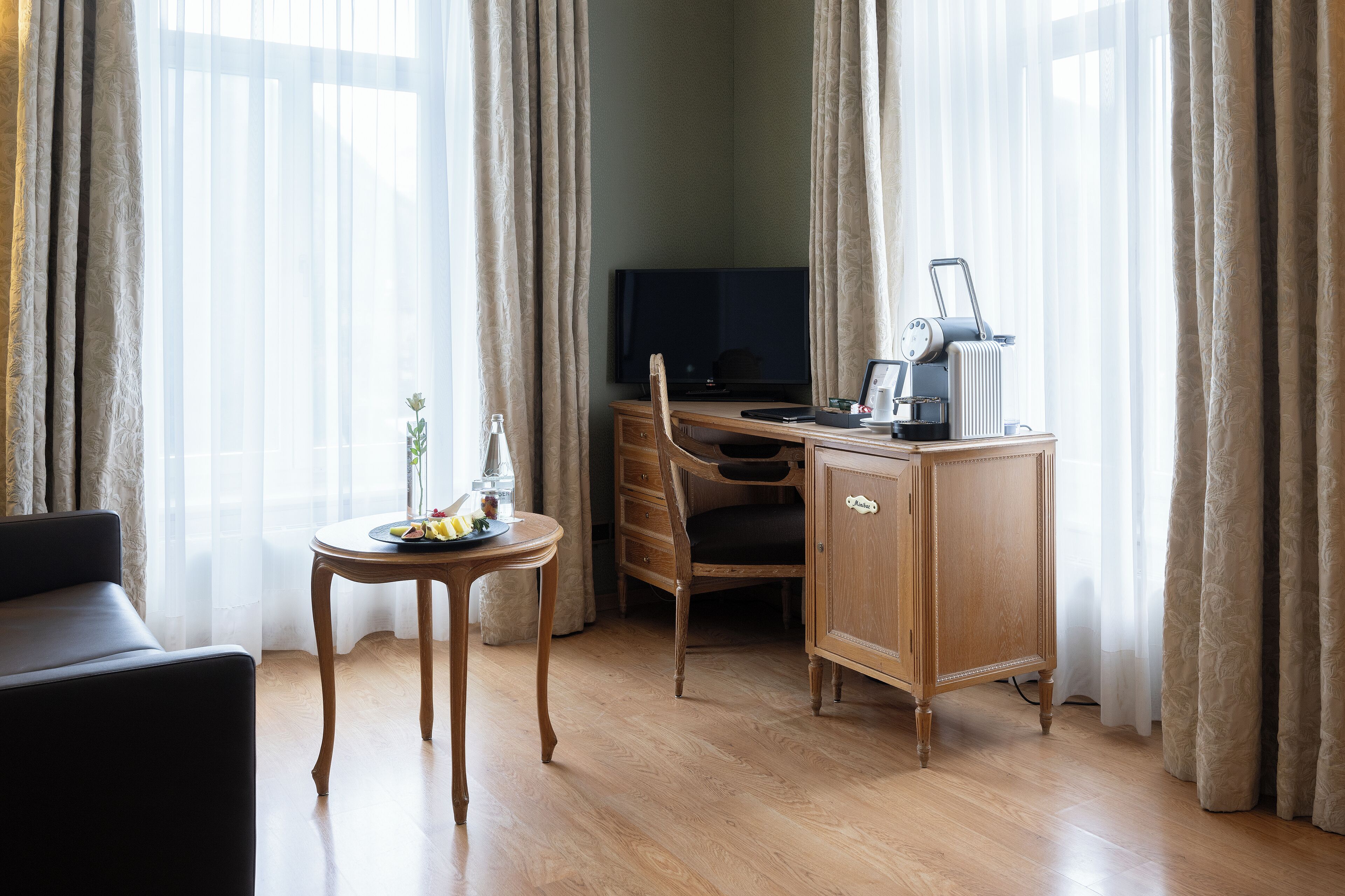 A hotel room with a wooden desk, a television, a radiator, a small table with a plant, and a sofa.