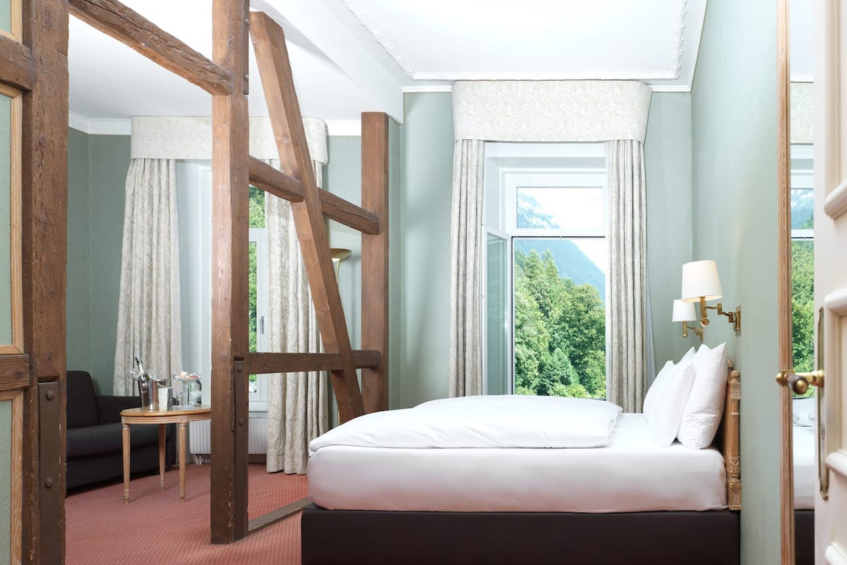 Ett hotellrum med en stor säng, en trästege, ett litet bord med en flaska och glas, och utsikt över grönskan genom fönstret.