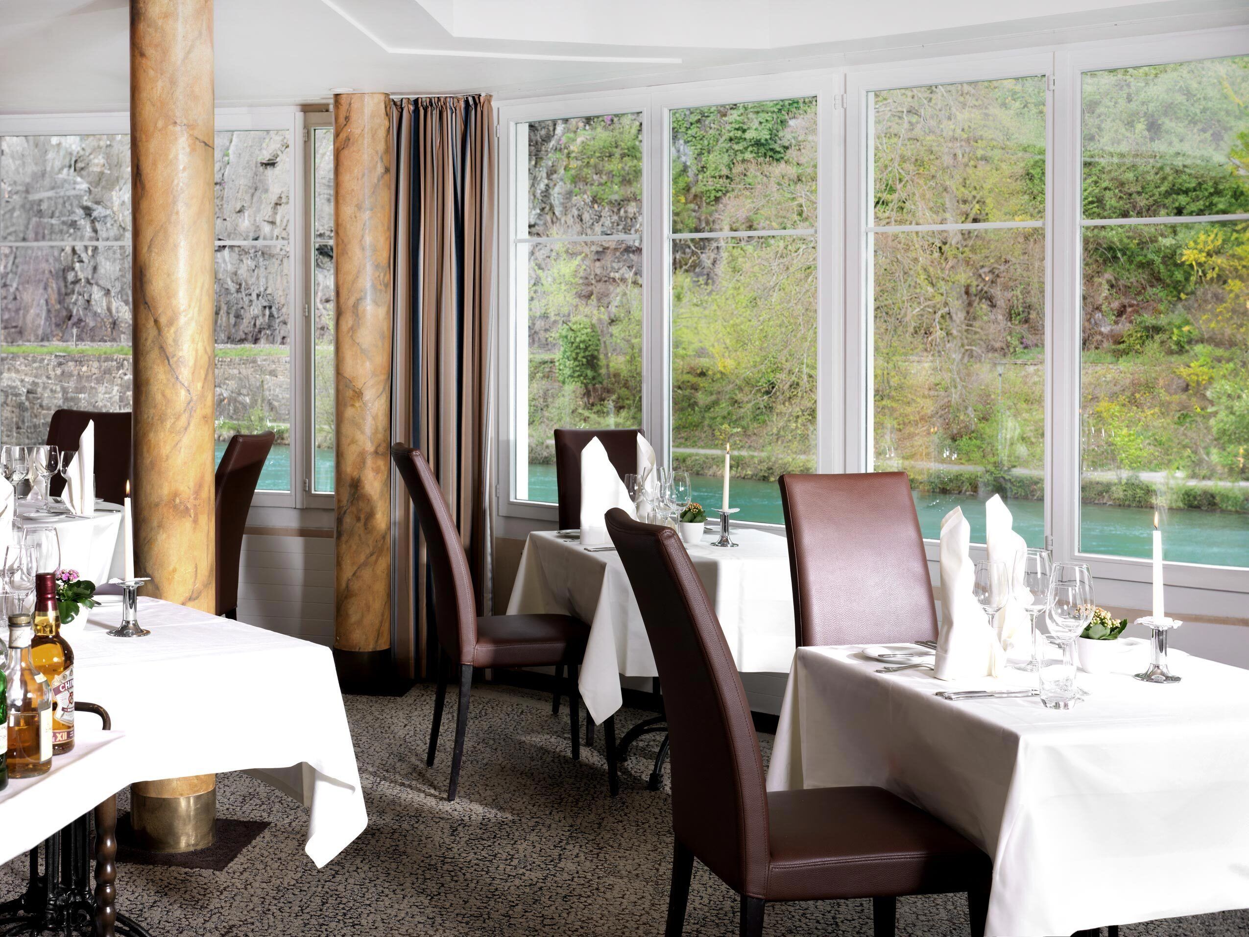 A dining area with tables set for two, large windows with a view of greenery and a body of water, and a marble column.