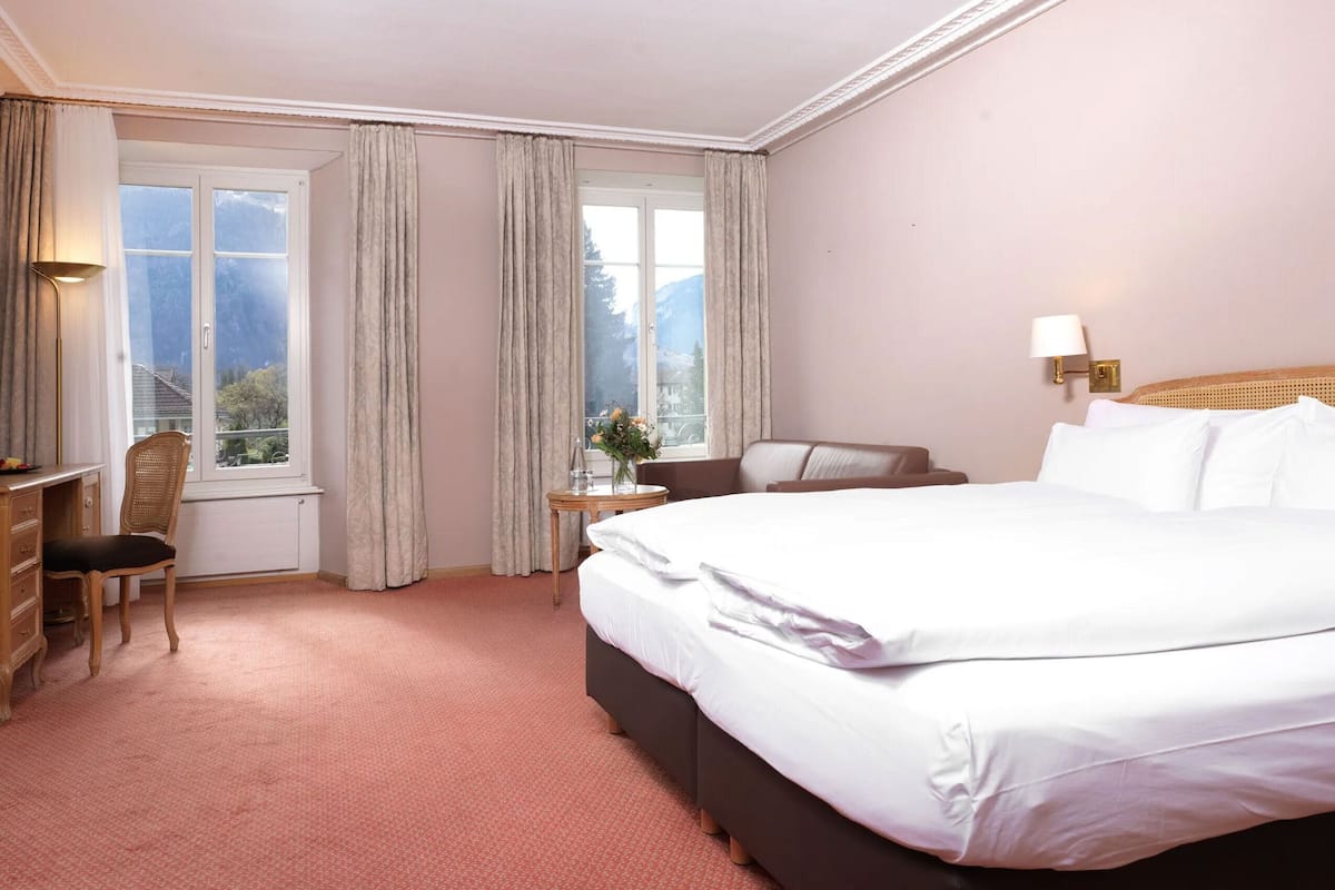 Ett hotellrum med en stor säng, ett skrivbord med en stol, ett litet bord med en vas och ett fönster med gardiner.