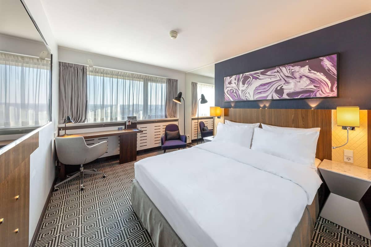 A hotel room with a large bed, a desk, a chair, and a view of the city.