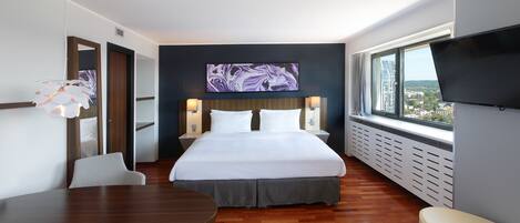 Habitación de hotel moderna con una cama grande, un televisor en la pared, una mesa de comedor y vistas a la ciudad desde la ventana.