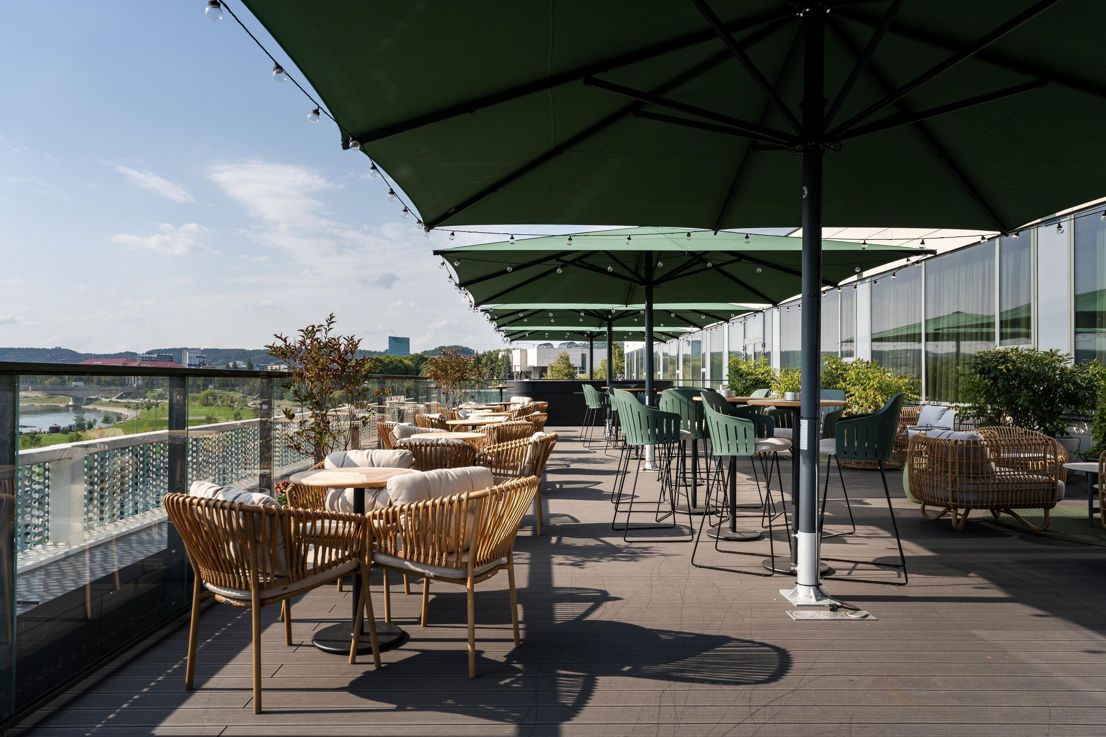 Una terraza en la azotea con mobiliario de mimbre, sombrillas verdes y vistas a la ciudad.