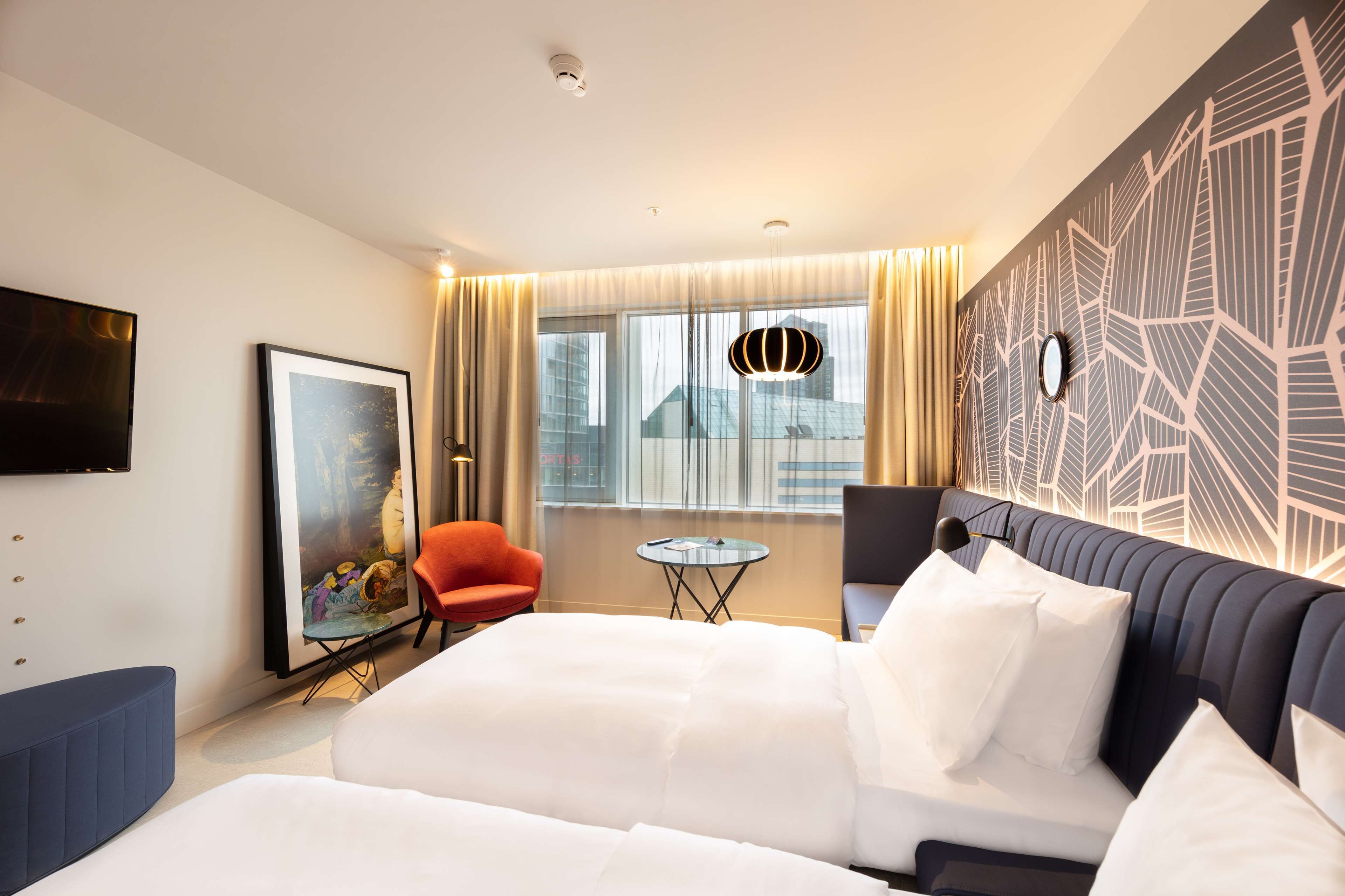 A hotel room with a large bed, a small table, a red chair, a TV, and a patterned wall.
