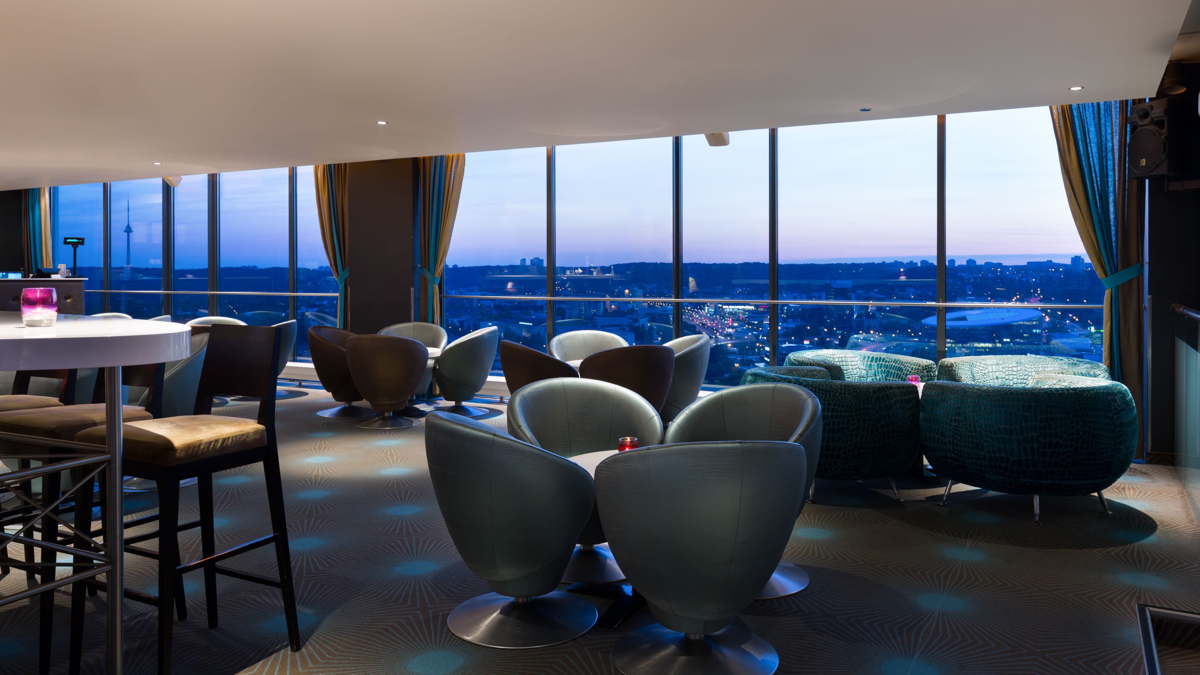 A modern hotel room with a panoramic city view, contemporary furniture, and a large window with curtains.
