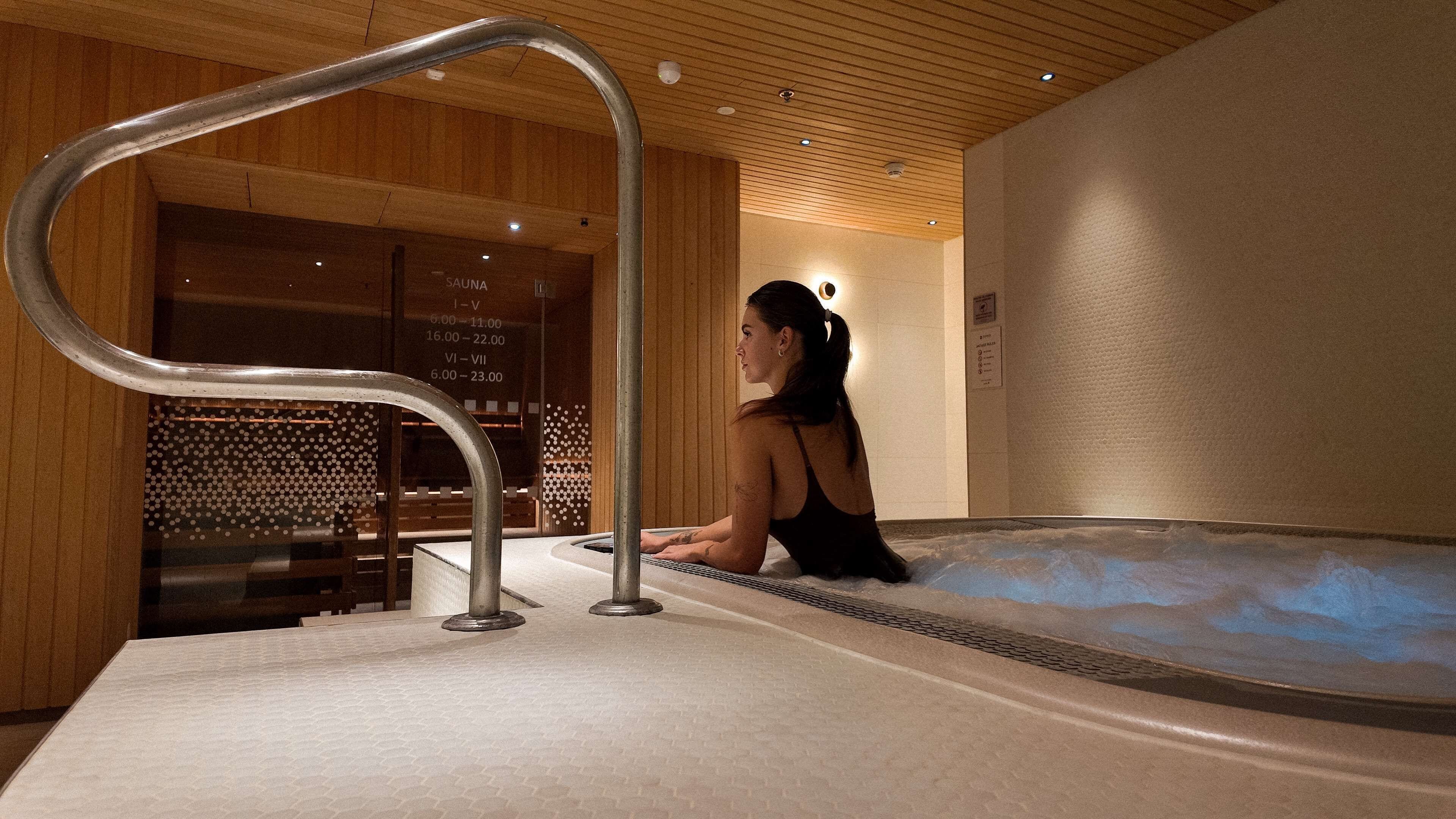 A woman sitting in a hot tub with a wooden interior and a visible sauna schedule on the wall.