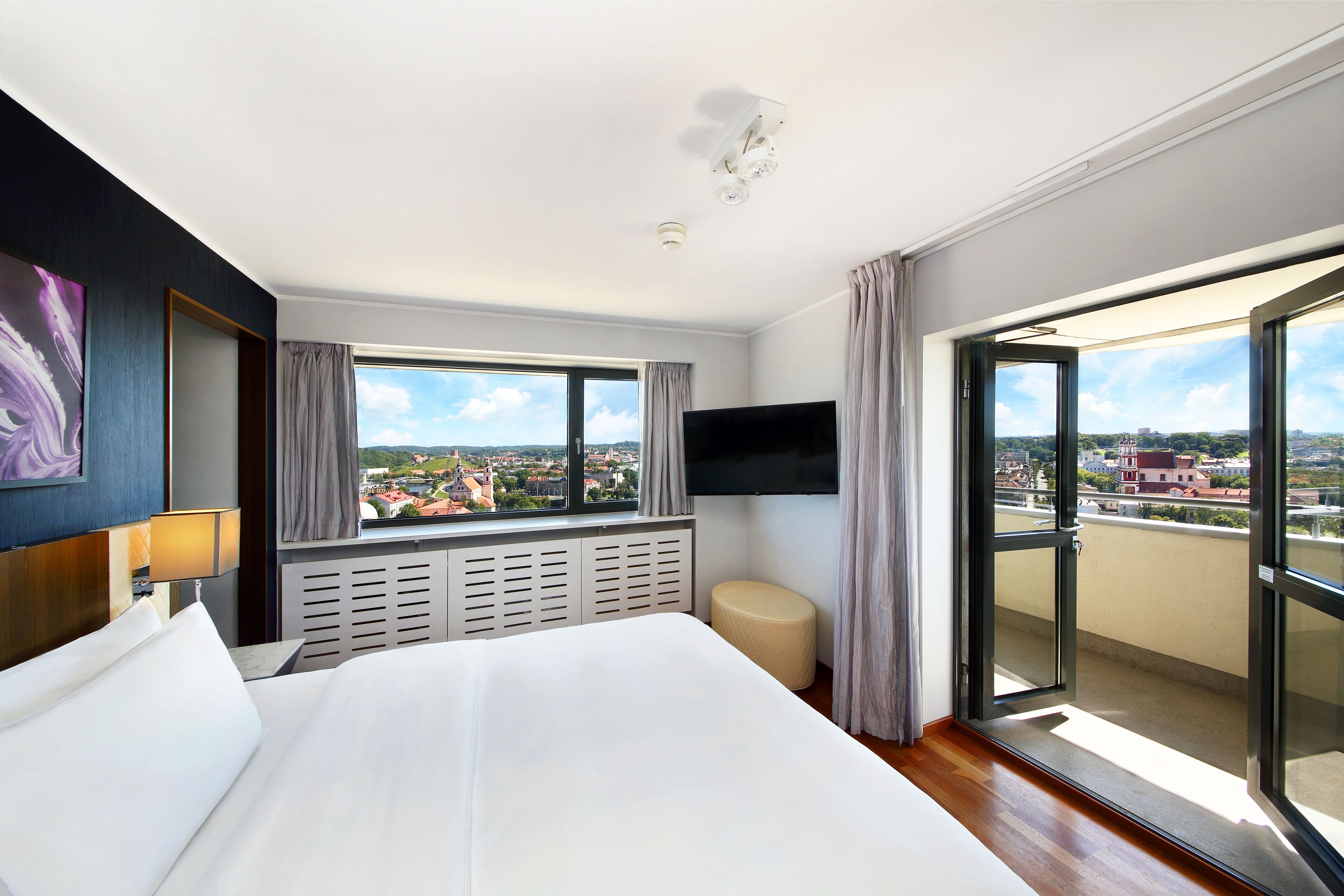 A hotel room with a large bed, a TV mounted on the wall, a window with a city view, and a balcony with sliding doors.