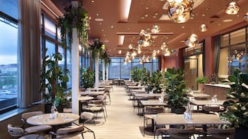 A spacious indoor dining area with multiple tables and chairs, large windows, and hanging spherical light fixtures.
