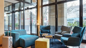 Un moderno vestíbulo de hotel con ventanas amplias, una zona de estar con sillones tapizados en azul y gris, un otoman amarillo y una maleta plateada.