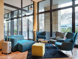 Hall moderna di un hotel con ampie finestre, un'area salotto con poltrone imbottite blu e grigie, un poggiapiedi giallo e una valigia argentata.