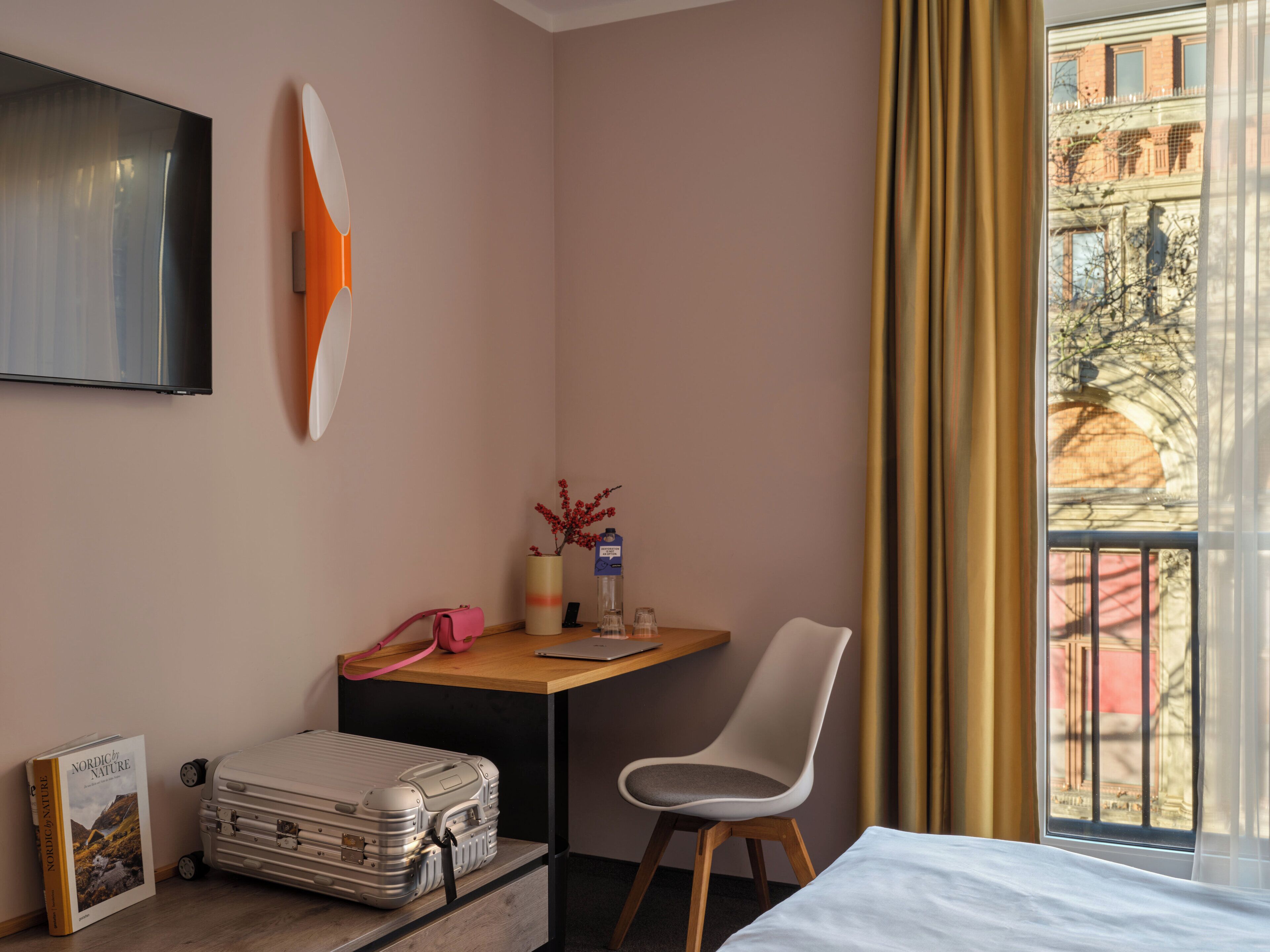 A hotel room with a bed, a desk with a chair, a suitcase, a TV mounted on the wall, and a window with curtains.