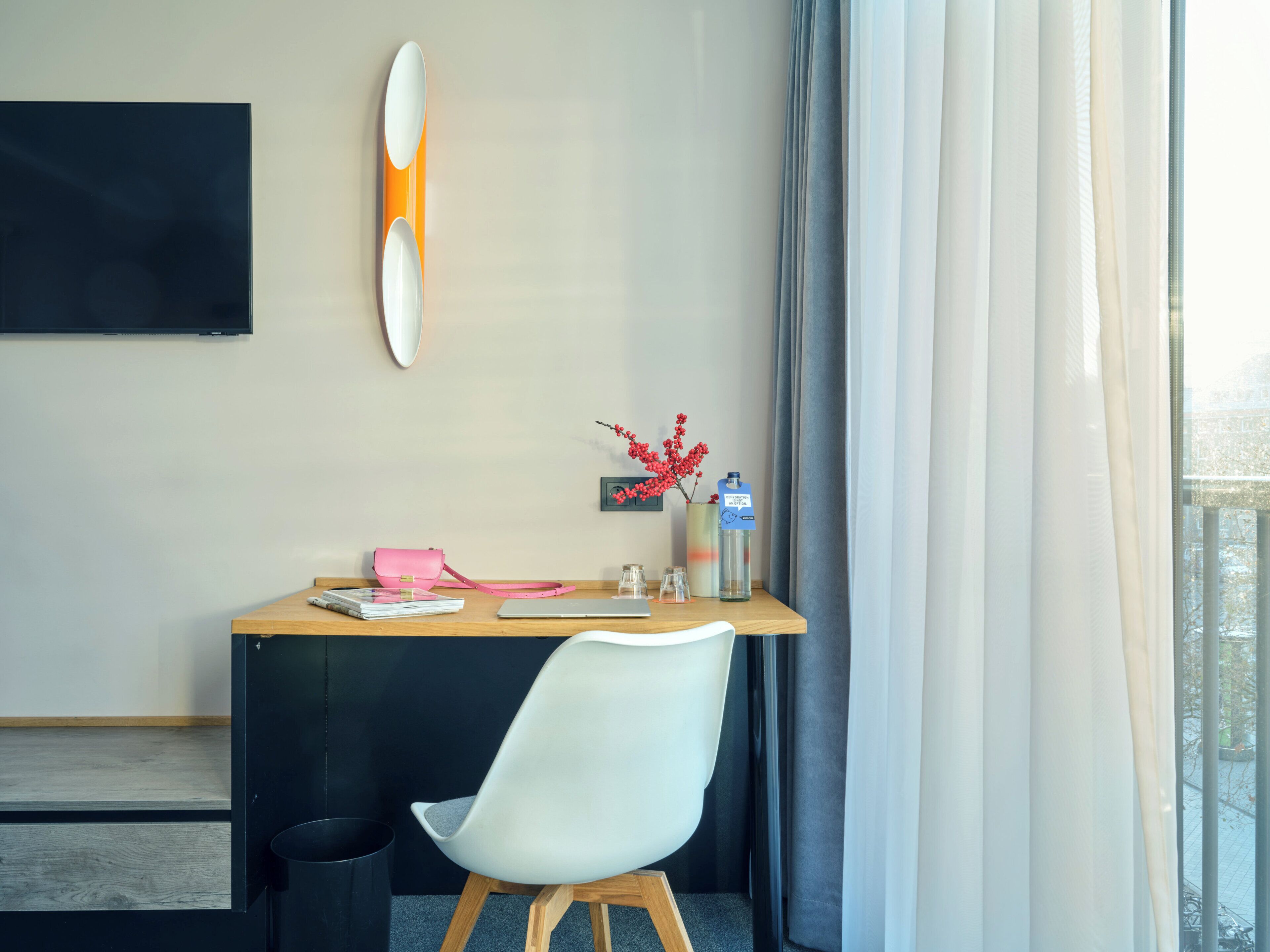 Habitación de hotel con un escritorio de madera, una silla blanca, un bolso rosa, una botella de agua y una tabla de surf en la pared.
