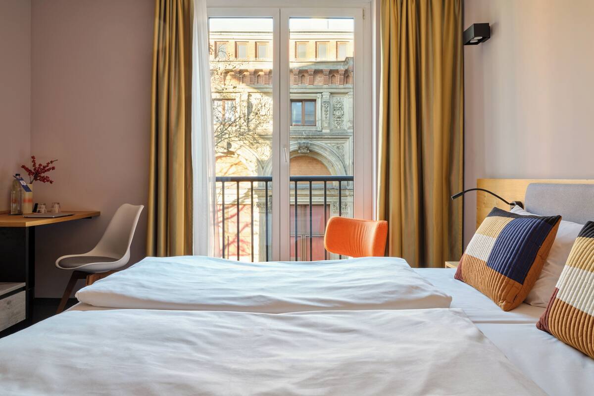 A hotel room with a bed, a desk, a chair, and a view of a building through the window.