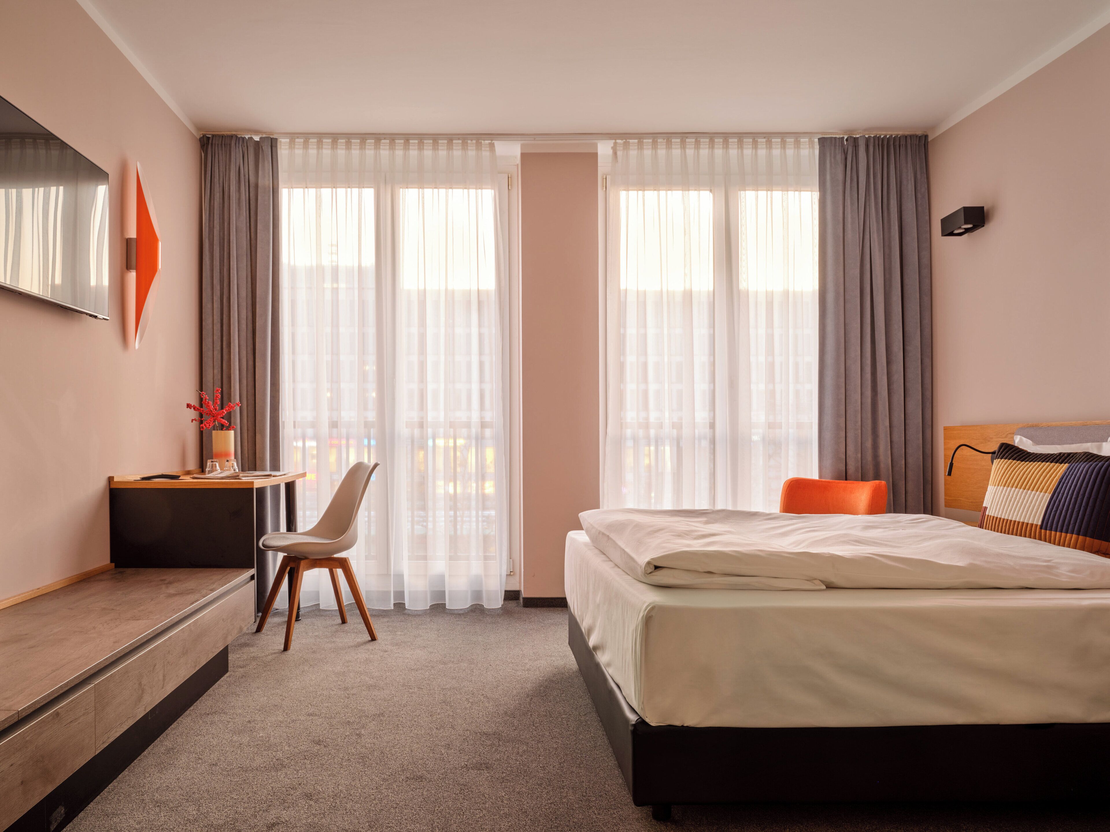A hotel room with a bed, a desk, a chair, and a large window with curtains.