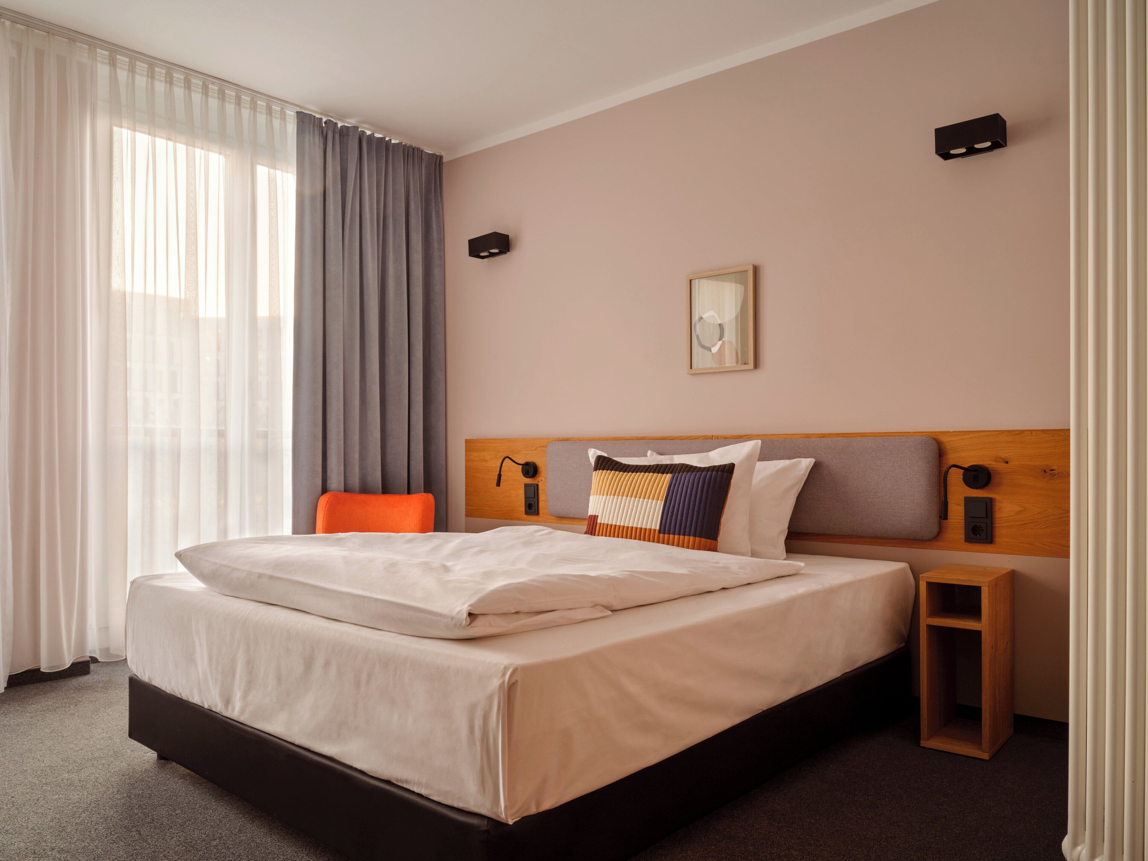 A hotel room with a large bed, a wooden headboard, a nightstand, a chair, and a window with curtains.