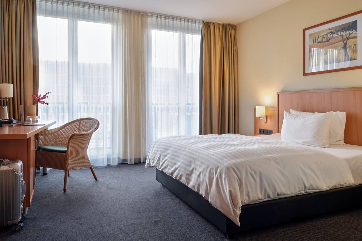 A hotel room with a bed, a desk, a chair, a lamp, and a painting on the wall.